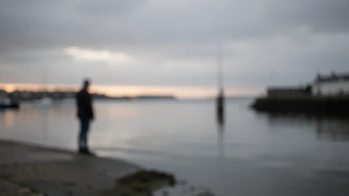 Generic image of a solemn dawn scene by the water, with a lone figure and overcast skies.